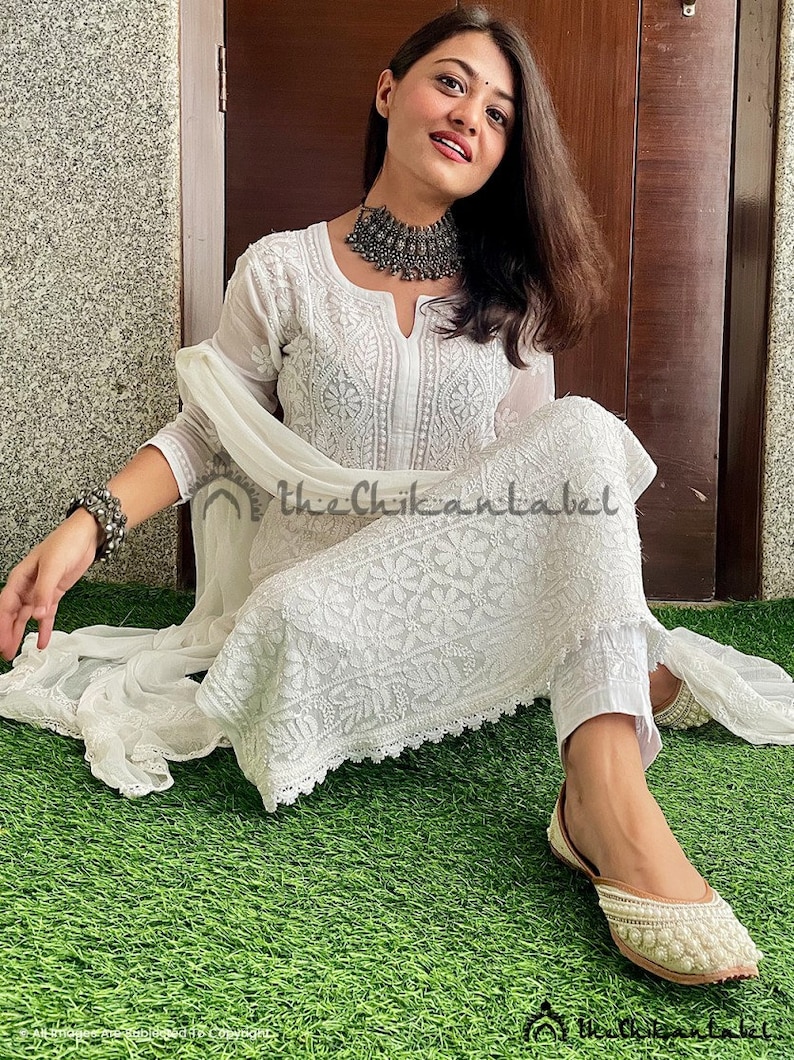 May include: A white chikankari kurta set featuring intricate embroidery and a delicate lace trim. The set includes a long-sleeved kurta, matching trousers, and a sheer dupatta. The model is wearing a silver necklace and bangles.