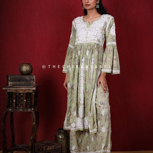 May include: A light green, long-sleeved, floral print kurta with white embroidery and bell sleeves. The kurta is paired with matching palazzo pants. The model is wearing silver earrings and silver shoes. The background is a solid red.