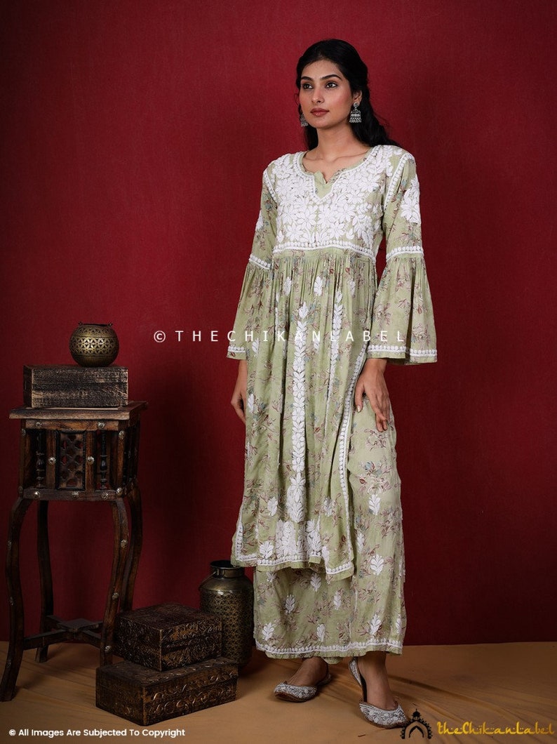 May include: A light green, long-sleeved, floral print kurta with white embroidery and bell sleeves. The kurta is paired with matching palazzo pants. The model is wearing silver earrings and silver shoes. The background is a solid red.