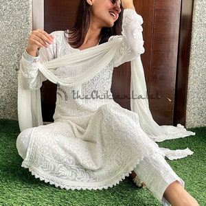 May include: A white cotton dress with intricate embroidery and a matching white dupatta. The dress has a round neckline and long sleeves. The dupatta is draped over the shoulders and is held in place by the hands. The dress is worn by a person sitting on a green grassy surface. The text "the Chikankari Label" is visible in the image.