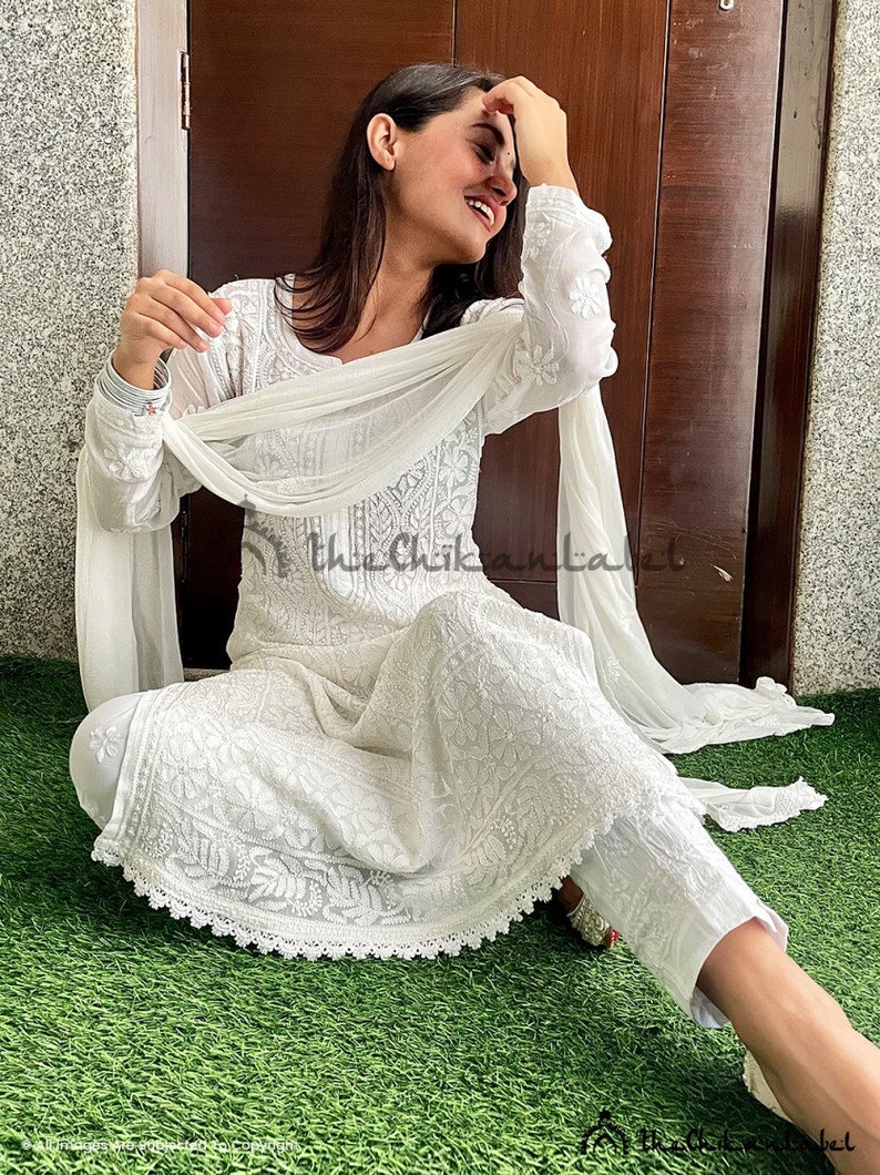 May include: A white cotton dress with intricate embroidery and a matching white dupatta. The dress has a round neckline and long sleeves. The dupatta is draped over the shoulders and is held in place by the hands. The dress is worn by a person sitting on a green grassy surface. The text "the Chikankari Label" is visible in the image.