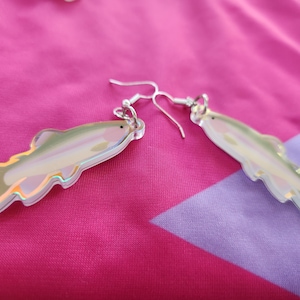 May include: A pair of iridescent fish-shaped earrings with silver hooks. The earrings are made of acrylic and have a holographic finish.