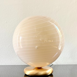 May include: A spherical table lamp with a cream glass globe and white horizontal stripes. The lamp has a round, gold-coloured base. This decorative lighting fixture is suitable for a bedside table or desk.