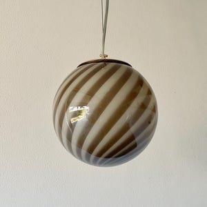 Peut inclure: Une suspension sphérique composée d'un globe en verre orné de rayures diagonales alternées en brun translucide et blanc opaque. Le globe est suspendu par un fil fin et fixé à un capuchon métallique circulaire sur la partie supérieure.