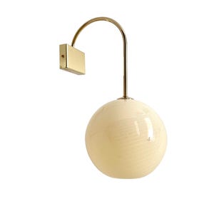 May include: A modern wall sconce featuring a spherical, cream-colored glass shade with subtle horizontal lines. The shade hangs from a curved, gold-toned metal arm connected to a rectangular wall mount.