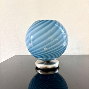 May include: A blue and white swirled glass orb vase. The vase has a round shape and sits on a silver-coloured base. The glass has a glossy finish and reflects light. The vase is a decorative item.