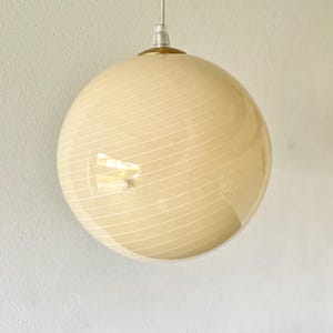 May include: A spherical pendant light in cream with a spiral of white lines. The light fitting hangs from a silver cable and a gold-coloured metal cap. The design is modern and the light is approximately 30 cm in diameter.