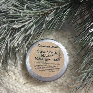 Puede incluir: Una pequeña lata redonda de metal de Seventeen Scents "Love Your Balls" Ball Butter. La etiqueta es de color canela con texto negro y un gráfico de árbol. La lata se encuentra sobre una superficie blanca texturizada, con ramas de pino verdes y blancas en el fondo.