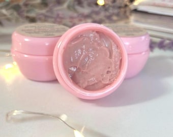 May include: Three small, pink cosmetic jars. One jar is open, revealing a pinkish-brown gel-like substance. The other two jars are closed, with a label on top. The jars are on a white surface, with string lights and blurred background elements.
