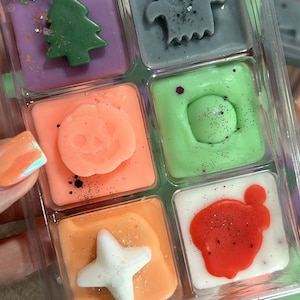 May include: A clear plastic clamshell container holding six square wax melts in various colours. The wax shapes include a green Christmas tree, a grey ghost, an orange jack-o'-lantern, a green circle, a white star, and a white square with red wax.