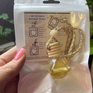 May include: A car air freshener in a clear plastic package. The product includes a small, geometric-shaped glass bottle filled with a yellow liquid, a wooden cap, and a braided cord. The package has instructions printed on it.
