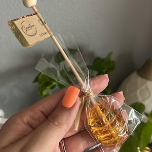 May include: A clear cellophane-wrapped lollipop with a wooden stick. The amber-colored candy is tied with twine. A small flag with the text "Seventeen Gourmet" is attached to the stick with a wooden ball on top.