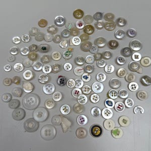 May include: A selection of buttons in a range of sizes and colours, including white, cream, and grey. Some buttons have decorative patterns and colourful accents. The buttons are made from various materials and have a pearlescent sheen.