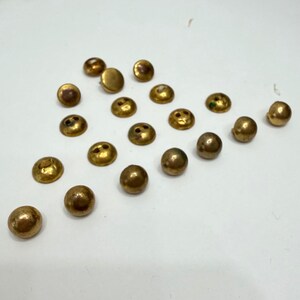 May include: A selection of small, round, antique brass buttons scattered on a white surface. The buttons have a slightly domed shape with two or three small holes for sewing. The brass has a warm, aged patina.