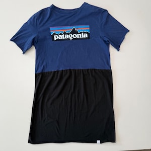 Op de afbeelding: Een tweekleurige t-shirtjurk. De bovenste helft is marineblauw met een rechthoekig logo met een bergketen en de merknaam "Patagonia". De onderste helft is effen zwart. De jurk heeft korte mouwen en een relaxte pasvorm.