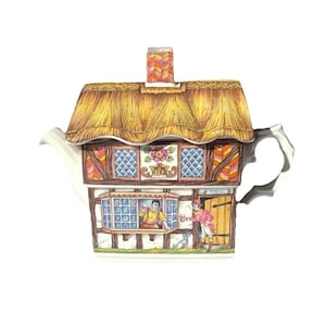 Puede incluir: Una tetera de cerámica con forma de cabaña con techo de paja, que presenta una escena pintada de una persona en una ventana y otra junto a una puerta de madera. El diseño incluye una chimenea, ventanas con patrón de diamantes azules y un perro.