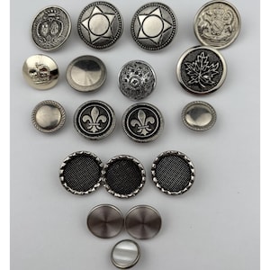 May include: A selection of silver-coloured buttons with diverse designs. The buttons display intricate details, such as stars, crowns, fleur-de-lis, and textured patterns. Some buttons have a raised relief design, while others are flat.
