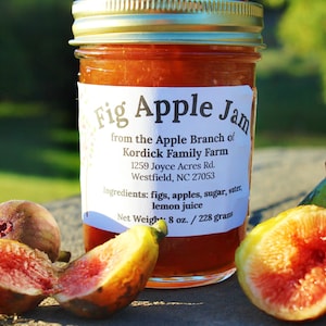 May include: A jar of Fig Apple Jam with a gold lid, labeled with the product name and farm details. Fresh figs, some sliced, are arranged around the jar. The jam is 8 oz. / 228 grams.