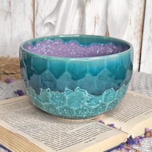 Peut inclure: Un bol en céramique turquoise avec un extérieur texturé et un intérieur violet moucheté. Le bol présente un motif hexagonal et repose sur un livre ouvert. Le design du bol évoque les vagues de l'océan.
