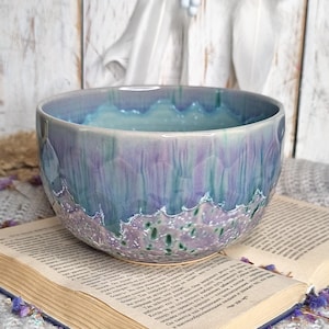 May include: A ceramic bowl with a textured, iridescent finish in shades of blue, purple, and green. The bowl is placed on an open book with aged pages. The bowl's rim has a decorative, scalloped edge.