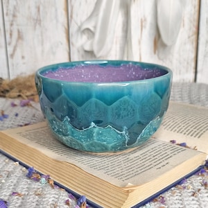 Puede incluir: Cuenco de cerámica artesanal con exterior turquesa texturizado e interior morado moteado. El cuenco está sobre un libro abierto con páginas envejecidas, sobre una superficie de punto gris. El diseño del cuenco presenta un patrón de ondas.