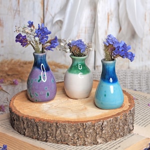 Peut inclure: Trois petits vases en céramique colorés avec des fleurs bleues séchées sont posés sur une tranche de bois. Les vases sont peints dans des tons de violet, de vert et de bleu. La tranche de bois repose sur un livre avec du texte visible.