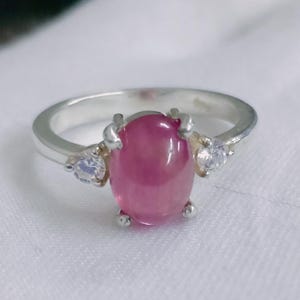 May include: A silver ring with a large, oval pink gemstone at its centre. Two smaller, clear gemstones are set either side of the main stone. The ring is displayed on a white surface.