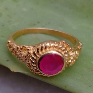 May include: A gold ring with a round, bright pink gemstone. The ring has an ornate, textured design around the gemstone and along the band. The ring is set against a green background.