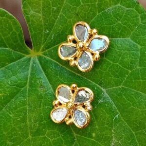 May include: A pair of gold flower-shaped earrings with clear gemstones. The earrings are on a green leaf.