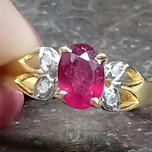 May include: A gold ring featuring a central oval-cut vibrant red gemstone held by four prongs. The band includes two small, clear, faceted stones on each side, set within silver-toned metal accents that contrast with the yellow gold band.