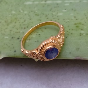 Pode incluir: Um anel de ouro com uma pedra preciosa azul. O anel tem um design ornamentado e texturizado ao redor da pedra preciosa e ao longo da banda. O anel est&aacute; sobre um fundo verde.