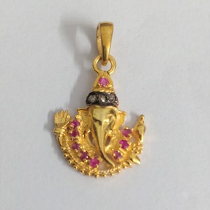 May include: A gold-colored pendant in the shape of Ganesha, adorned with small red gemstones and a black and silver accent. The pendant features intricate details and a loop for a chain.