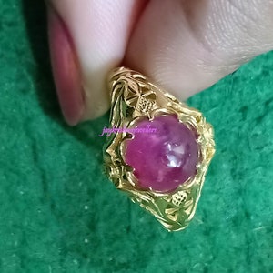 以下が含まれることがあります： 大きな楕円形のピンク色の宝石が付いた金の指輪。指輪には、宝石の周りとバンドに華やかな花の模様があります。指輪は緑色の背景にあります。