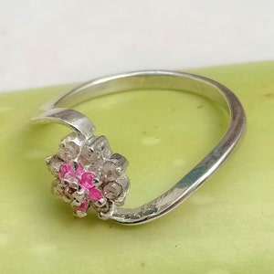 May include: A silver ring with a floral design. The flower has pink and clear gemstones. The ring band curves around the finger, with the flower design on the top. The ring is set against a light green background.
