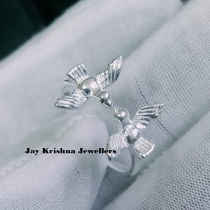 Op de afbeelding: Een zilverkleurige open ring met twee vogels die naar elkaar toe kijken met gespreide vleugels. Het metalen oppervlak heeft een gepolijste afwerking met gestructureerde vleugeldetails. De tekst "Jay Krishna Jewellers" staat in het zwart vermeld.