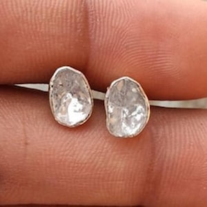 Puede incluir: Un par de pendientes de diamantes transparentes de forma irregular. Los diamantes están engastados en un metal de color plateado. Los pendientes se muestran sobre un fondo color piel.