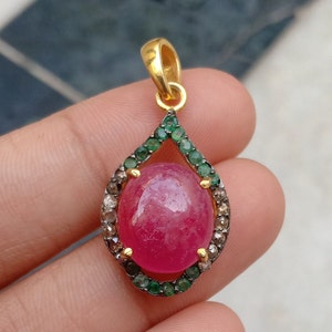 May include: A teardrop-shaped pendant with a large, oval, pink gemstone at its center. The gemstone is surrounded by a border of small, green gemstones and a ring of smaller, clear gemstones. The pendant has a gold-colored bail.