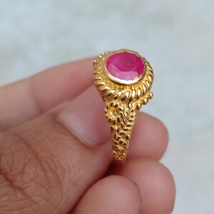 Könnte beinhalten: Ein goldener Ring mit einem großen, ovalen Rubin-Edelstein. Der Ring hat ein detailliertes, strukturiertes Band.