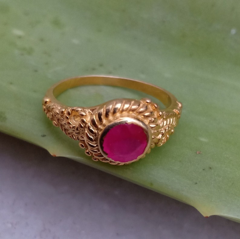 May include: A gold ring with a round, bright pink gemstone. The ring has an ornate, textured design around the gemstone and along the band. The ring is set against a green background.