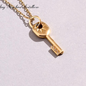 May include: Gold-colored key pendant necklace. The key pendant has a sunburst design and is adorned with small, sparkling stones. The necklace chain is also gold-colored. The text "Jay Krishna Jewellers" is visible at the top.