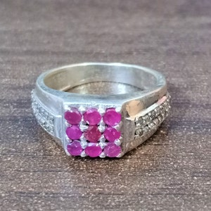 Peut inclure: Une bague en argent avec un groupe carré de neuf pierres précieuses rouges au centre. La bande comporte une rangée de pierres plus petites et claires de chaque côté du carré central. La bague est posée sur une surface marron.