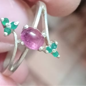 May include: A silver ring featuring a large, oval-cut, pink gemstone and small green gemstones. The ring has a unique, asymmetrical design with the gemstones set on delicate, curving branches.