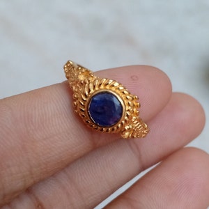 Pode incluir: Um anel de ouro com uma pedra preciosa redonda azul escura. O anel tem um design ornamentado e texturizado ao redor da gema e ao longo da banda. O anel &eacute; segurado em uma m&atilde;o, mostrando os detalhes intrincados da joia.