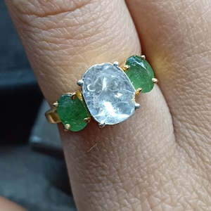 Puede incluir: Un anillo de oro con una gema clara grande de forma irregular flanqueada por dos gemas verdes ovaladas más pequeñas. El engaste es dorado con garras plateadas que sujetan la piedra central.