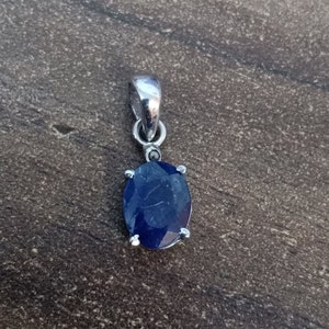 May include: A silver pendant with a large, oval, deep blue gemstone. The gemstone is secured by four silver prongs. The pendant has a silver bail for a chain. The background is a textured, dark grey surface.