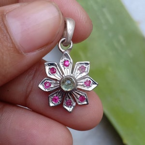 May include: Silver pendant with a flower design. The pendant features a central green gemstone surrounded by eight pink gemstones. The flower petals are silver and pointed. The pendant has a small loop at the top for a chain.