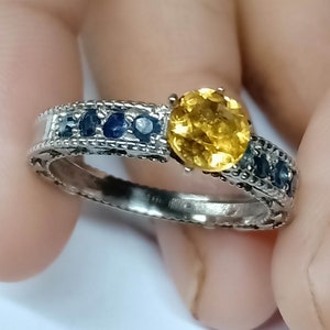 Peut inclure: Une bague en argent ornée d'une grosse pierre jaune ronde en son centre. La monture est ornée de petites pierres bleues foncées et de détails complexes. Un bijou classique et élégant.