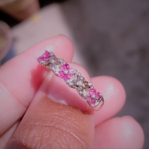 Peut inclure: Une bague en argent ornée de deux rangées de petites pierres précieuses rondes. Les pierres précieuses alternent entre le rose vif et le clair, créant un effet scintillant. La bague est tenue dans une main.