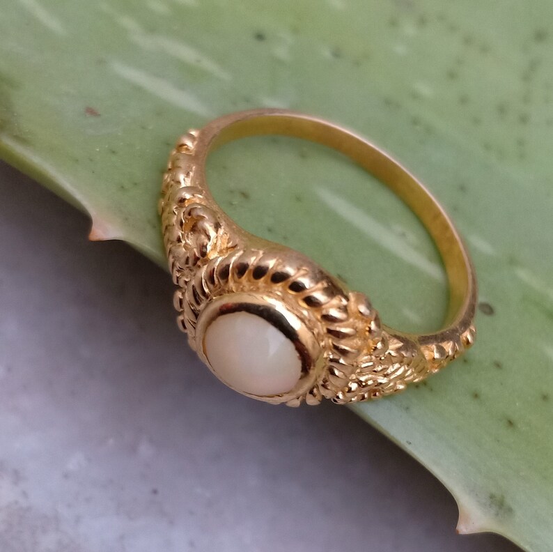 Pode incluir: Um anel dourado com uma pedra ovalada de cor creme. O anel tem um design ornamentado e texturizado ao redor da pedra e ao longo da banda. O anel est&aacute; sobre um fundo verde.