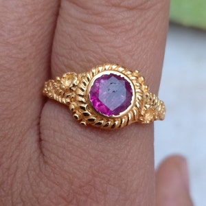 Pode incluir: Um anel de ouro com uma grande pedra preciosa magenta redonda. O anel tem uma banda detalhada e texturizada com desenhos florais e semelhantes a cordas. A pedra &eacute; definida em uma configura&ccedil;&atilde;o de moldura.